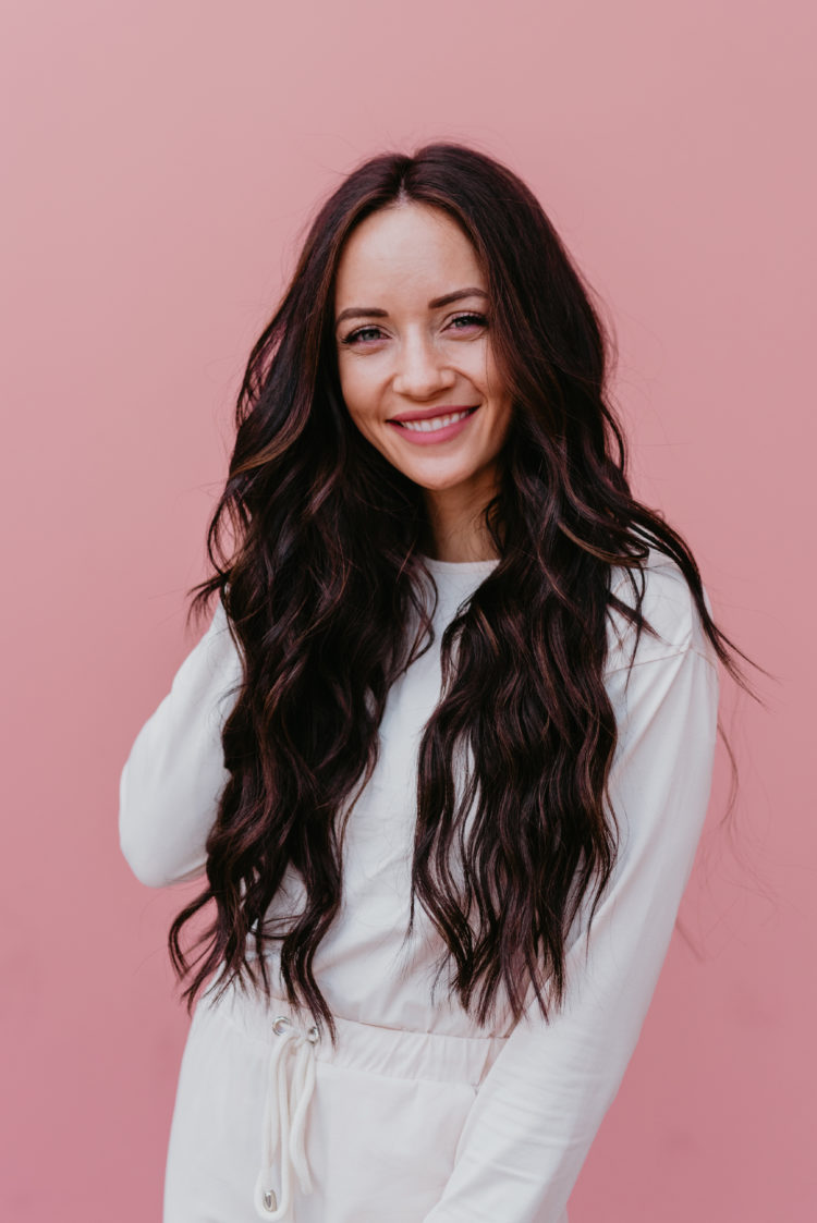 Natural Beaded Extensions by popular Las Vegas beauty blog, Outfits and Outings: image of a woman with long brown wavy hair and standing in front of a pink wall while wearing a white jump suit.