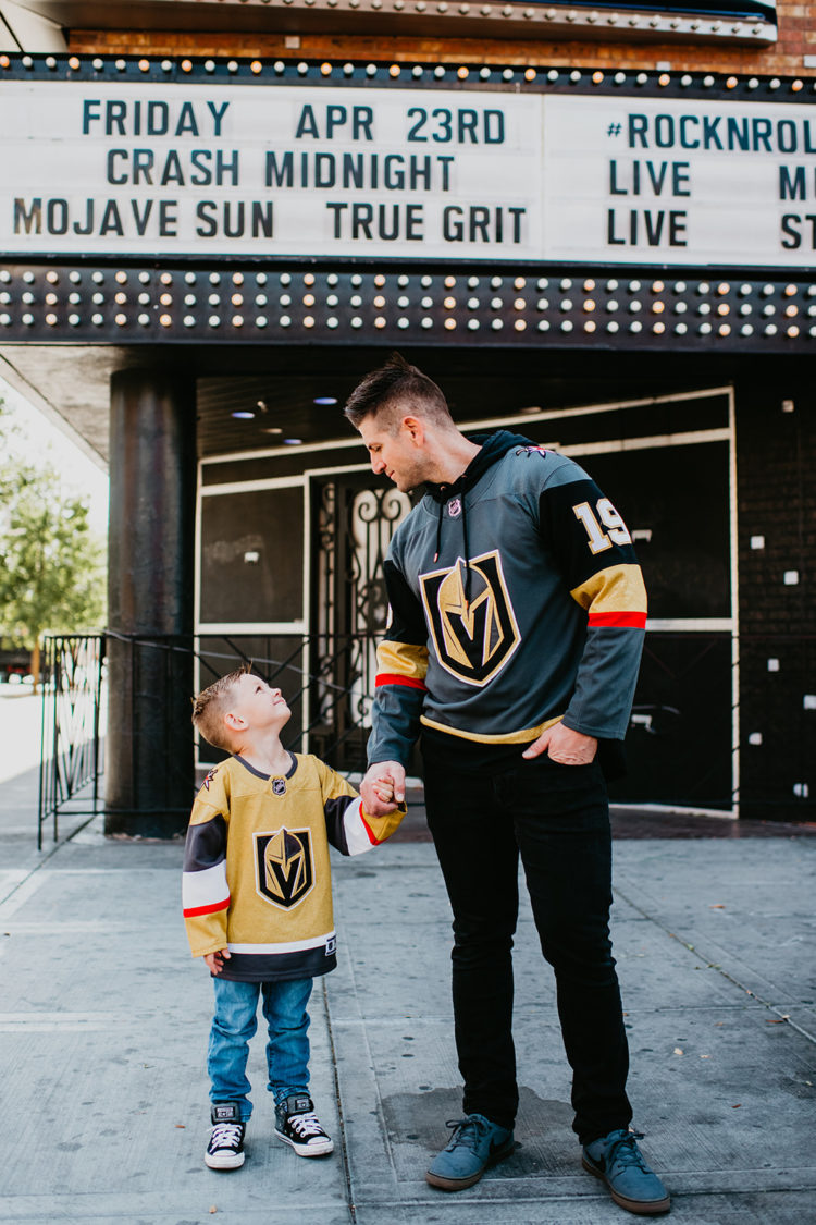 Golden Knights Gear by popular Las Vegas fashion blog, Outfits and Outings: image of a dad and his young son holding hands and standing under a movie theater marquee and wearing a Golden Knights sweatshirt hoodie and Golden Knights jersey. 