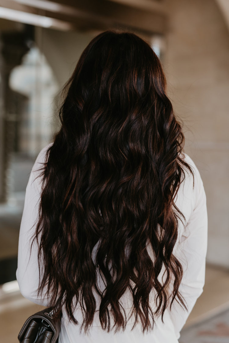 Natural Beaded Extensions by popular Las Vegas beauty blog, Outfits and Outings: image of a woman with long brown wavy hair and wearing a white jump suit.