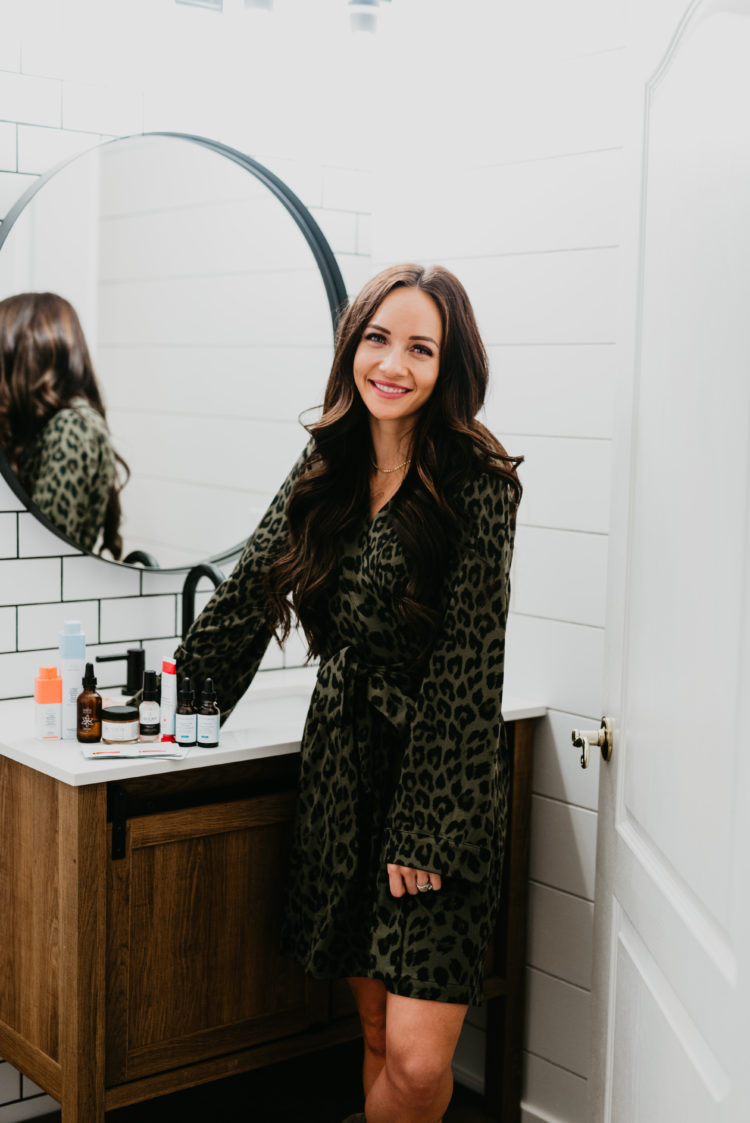 Acne and Anti Aging Skincare Routine by popular Las Vegas beauty blog, Outfits and Outings: image of a woman wearing a green and black leopard print robe and standing next to various acne and skincare products resting on a bathroom vanity. 