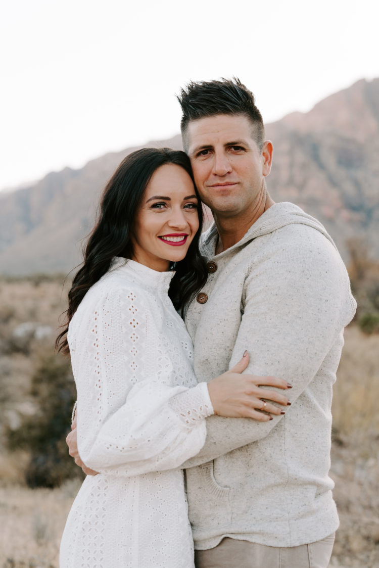Family Picture Ideas by popular Las Vegas fashion blog, Outfits and Outings: image of a man and woman embracing each other in the desert and wearing a white eyelet dress, cream hooded pullover, and tan pants. 
