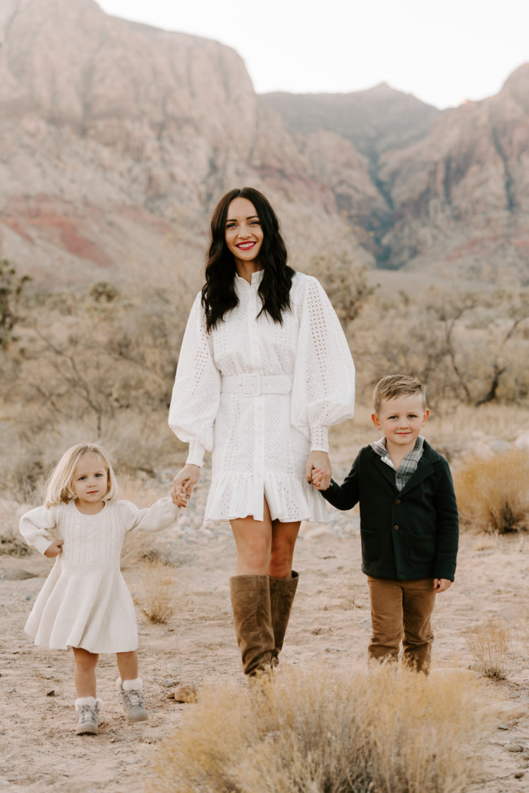 Family Picture Ideas by popular Las Vegas fashion blog, Outfits and Outings: image of a mom and her two kids holding hands in the desert and wearing a white eyelet dress, brown suede boots, cream dress, brown pants, and green sweater. 