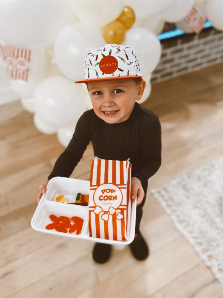 Birthday Party Ideas by popular Las Vegas lifestyle blog, Outfits and Outings: image of a little boy wearing a soda print cap and black pajama set and holding a white plastic food tray filled with red Swedish fish, gummy bears, and a red and white stripe popcorn bag.  