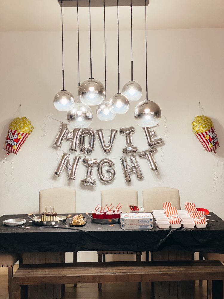 Birthday Party Ideas by popular Las Vegas lifestyle blog, Outfits and Outings: image of a movie night balloon garland hanging behind a table set with a cake, metal tray filled with red plastic cups filled with soda, pizza boxes, and white plastic food trays filled with gummy candies and red and white stripe popcorn bags. 