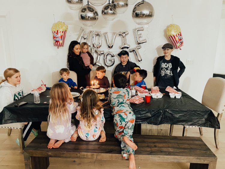 Birthday Party Ideas by popular Las Vegas lifestyle blog, Outfits and Outings: image of a group of kids wearing pajamas and standing around a table with a black table cloth and set with a birthday cake with lit candles and white food trays filled with popcorn and gummy candies. 