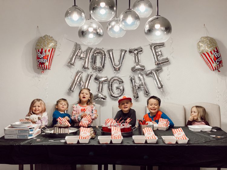 Birthday Party Ideas by popular Las Vegas lifestyle blog, Outfits and Outings: image of a group of kids wearing pajamas and standing around a table with a black table cloth and set with a birthday cake with lit candles and white food trays filled with popcorn and gummy candies. 