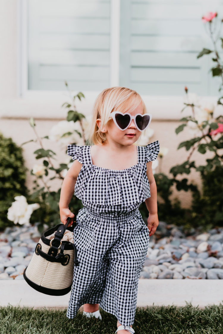 Easter Basket Ideas by popular Las Vegas life and style blog, Outfits and Outings: image of a little girl wearing a black and white gingham flutter sleeve romper, white heart frame sunglasses, and white sandals. 