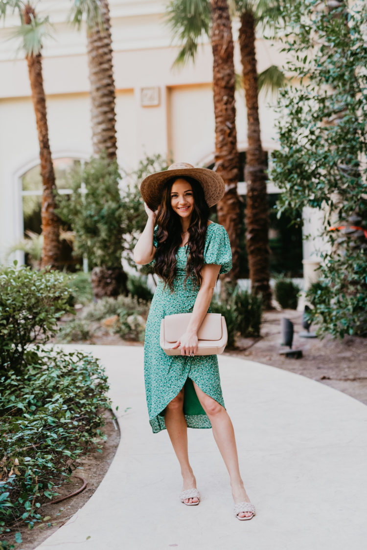 Wedding Guest Dresses by popular Las Vegas fashion blog, Outfits and Outings: image of a woman standing on a sidewalk near some palm trees and wearing a green floral print puff sleeve dress with a straw sun hat and tan braided strap block heel sandals while holding a tan purse. 