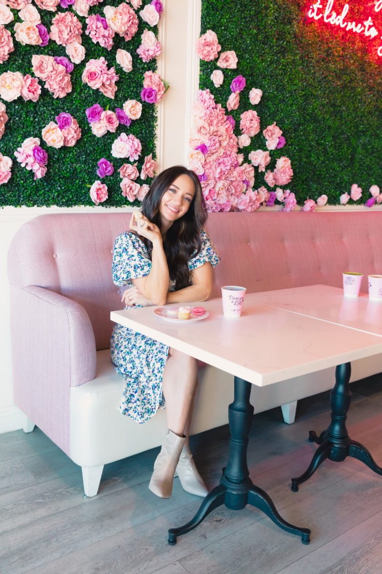 Wedding Guest Dresses by popular Las Vegas fashion blog, Outfits and Outings: image of a woman sitting at a table in front of al floral wall display and wearing a white and blue floral print dress and tan ankle boots. 