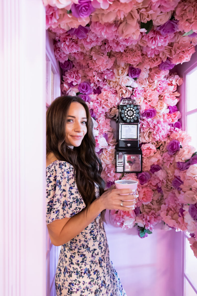 Wedding Guest Dresses by popular Las Vegas fashion blog, Outfits and Outings: image of a woman standing in a phone booth decorated with purple and pink flowers and wearing a white and blue floral print dress. 