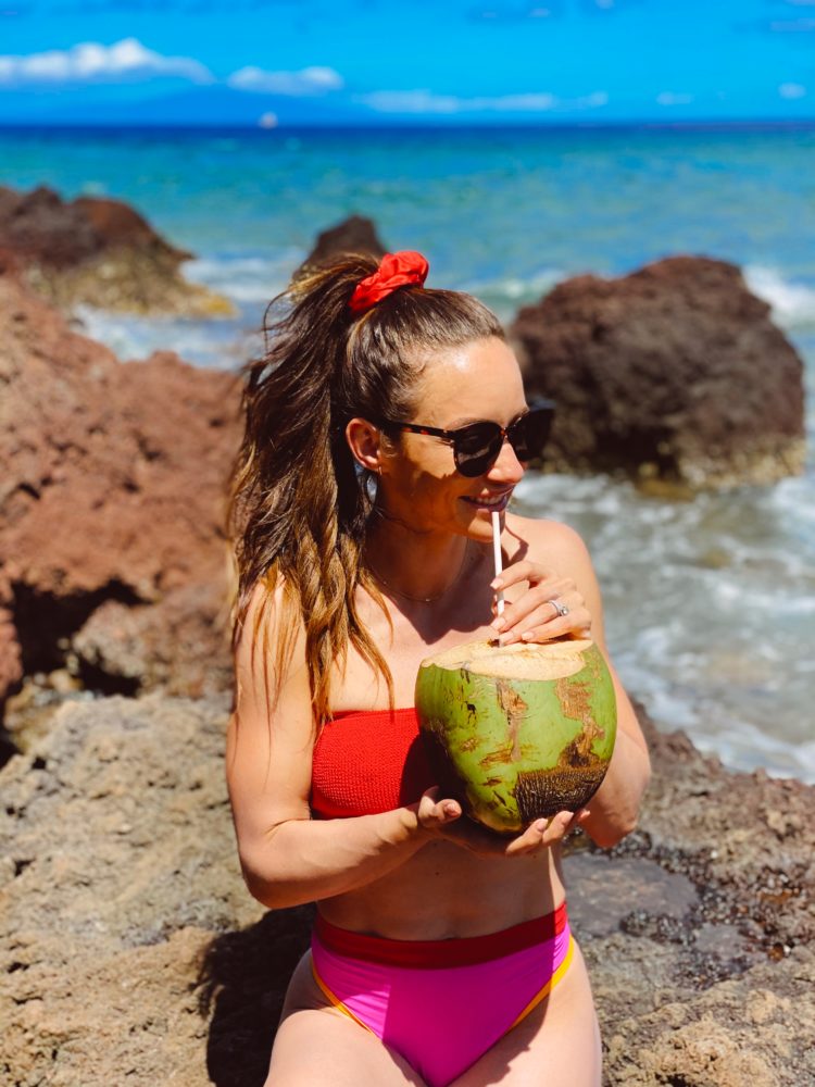 Best Restaurants in Maui by popular Las Vegas travel blog, Outfits and Outings: image of a woman wearing a pink and red two piece swimsuit white standing next to the ocean and drinking out of a coconut. 