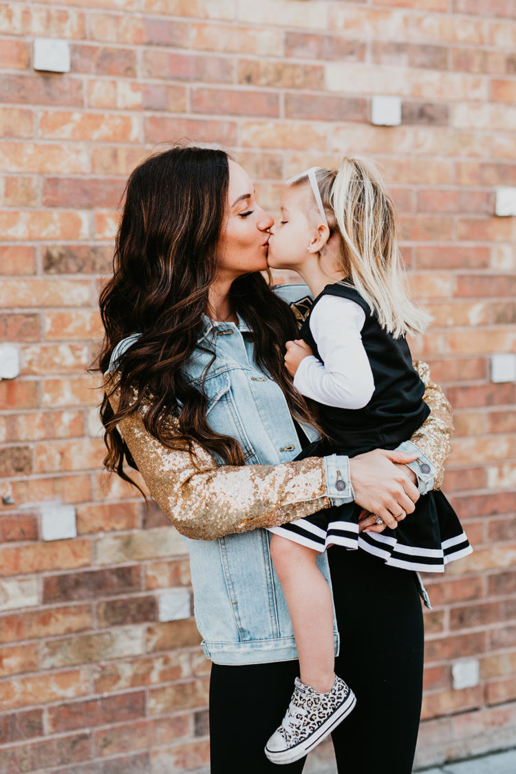 Golden Knights Gear by popular Las Vegas fashion blog, Outfits and Outings: image of a mom holding her young daughter and kissing her on the lips while wearing a gold sequin sleeve denim Golden Knights jacket and black Golden Knights cheerleading uniform and leopard print sneakers. 