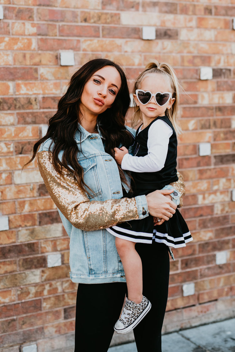 Golden Knights Gear by popular Las Vegas fashion blog, Outfits and Outings: image of a mom holding her young daughter and wearing a gold sequin sleeve denim Golden Knights jacket, black Golden Knights cheerleader uniform, heart frame sunglasses, and leopard print sneakers. 