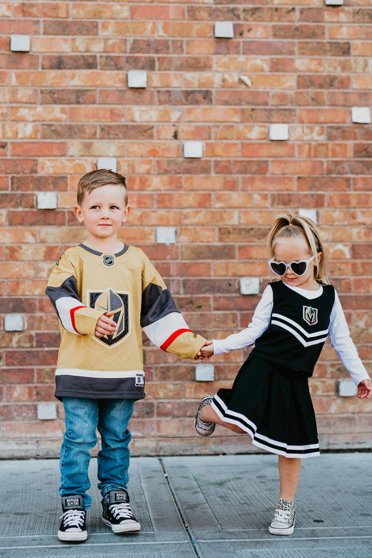 Golden Knights Gear by popular Las Vegas fashion blog, Outfits and Outings: image of a young boy and girl holding hands in front of a brick wall and wearing a Golden Knights jersey and Golden Knights cheerleading uniform. 