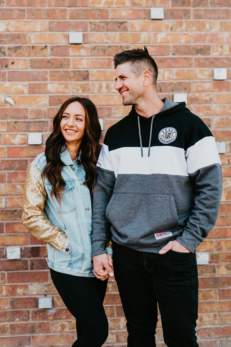 Golden Knights Gear by popular Las Vegas fashion blog, Outfits and Outings: image of a husband and wife standing together and holding hands in front of a brick wall and wearing a Golden Knights gold sequin sleeve denim jacket and Golden Knights sweatshirt hoodie. 