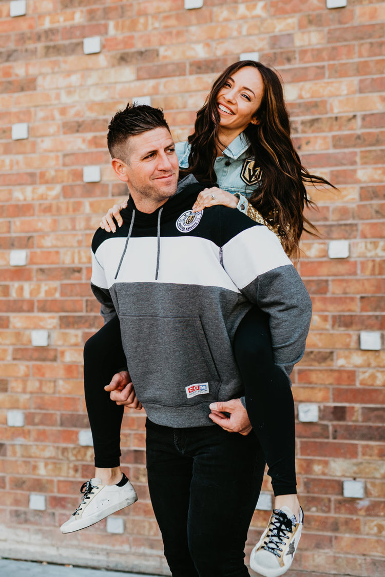Golden Knights Gear by popular Las Vegas fashion blog, Outfits and Outings: image of a woman on her husband's back and wearing Golden Goose sneakers, Golden Knights pullover sweatshirt hoodie, and Golden Knights gold sequin sleeve denim jacket. 