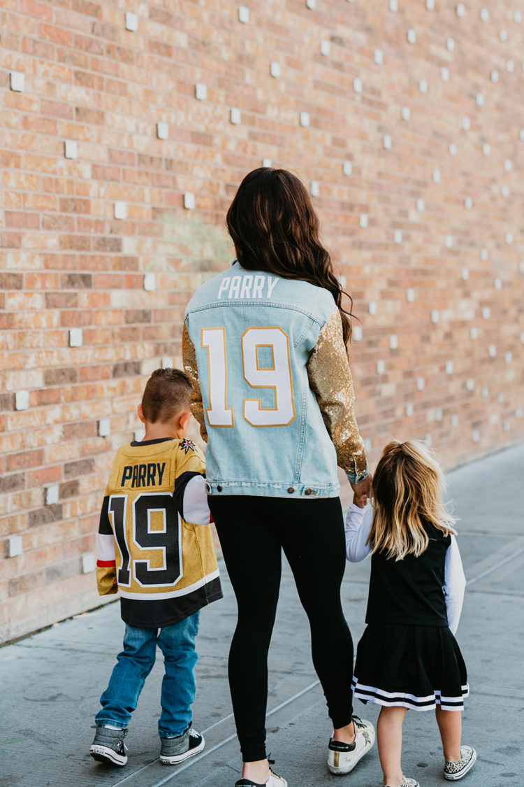 Golden Knights Gear by popular Las Vegas fashion blog, Outfits and Outings: image of a mom holding her young son and young daughters hands while they walk on a sidewalk and wear a Golden Knights jersey, Golden Knights cheerleading uniform, and Golden Knights gold sequin sleeve denim jacket. 