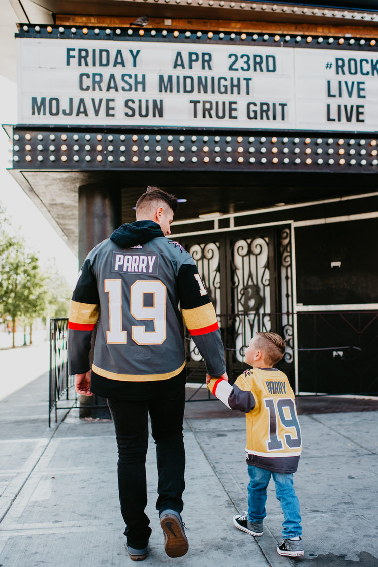 Golden Knights Gear by popular Las Vegas fashion blog, Outfits and Outings: image of a dad holding his young son's hand and walking under a movie theater marquee and wearing a leather Golden Knights jacket and Golden Knights jersey. 