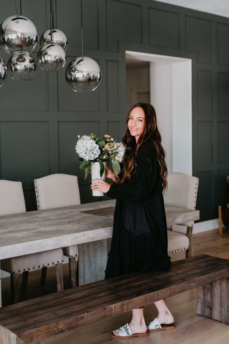 Copper and Tweed by popular Las Vegas life and style blog, Outfits and Outings: image of a woman wearing a black maxi dress and standing at a Copper and Tweed dining table while holding a vase of flowers.  