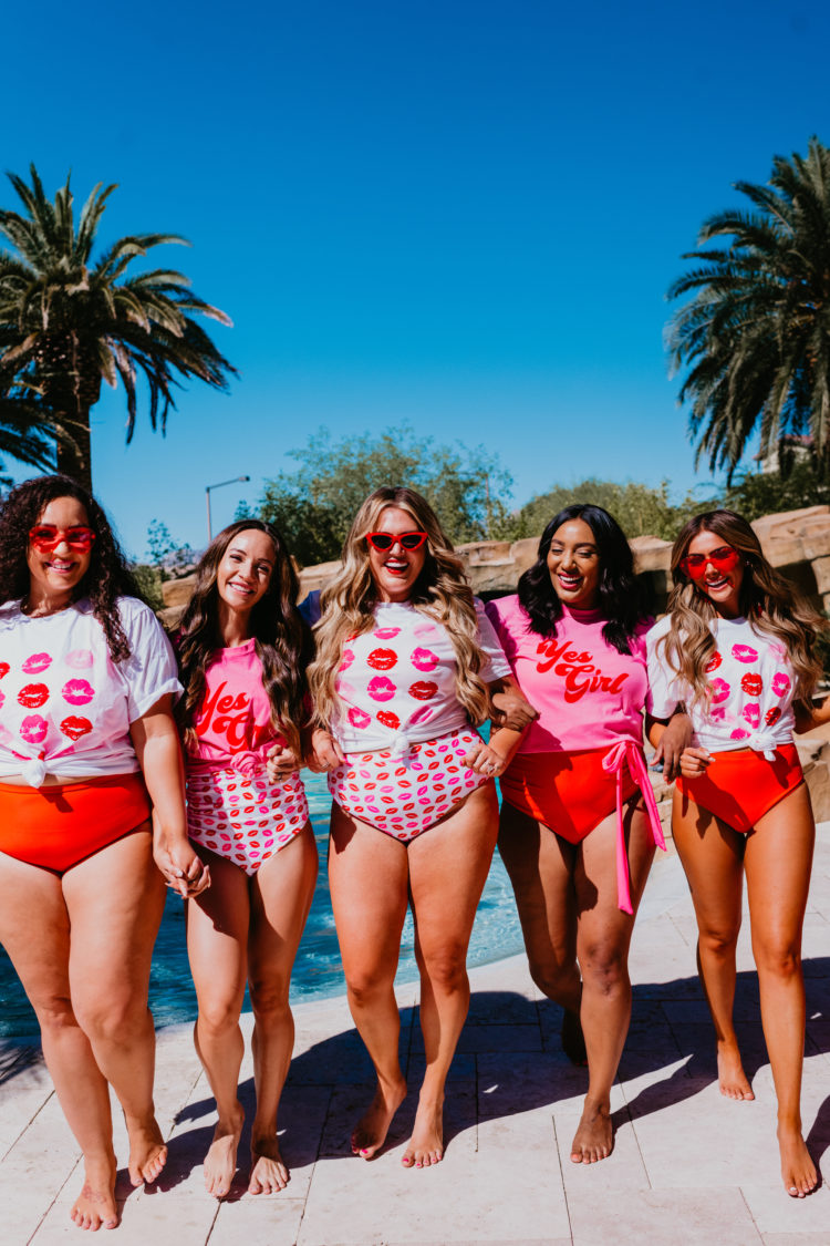 Sassy Red Lipstick x Pink Desert Swimwear featured by top Las Vegas fashion blogger, Outfits & Outings | Sassy Red Lipstick by popular Las Vegas fashion blog, Outfits and Outings: image of a group of women standing together outside by a swimming pool and wearing pink and red one piece and two piece swimsuits, lip print t-shirts, pink yes girl t-shirts, and red frame sunglasses.  