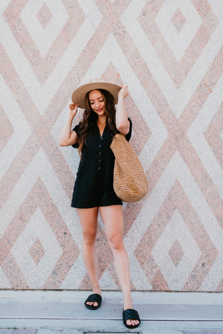 Cute Summer Rompers for Women by popular Las Vegas fashion blog, Outfits and Outings: image of a woman standing next to a wall mural and wearing a black romper with a straw fedora hat and black slide sandals and holding a straw tote bag. 