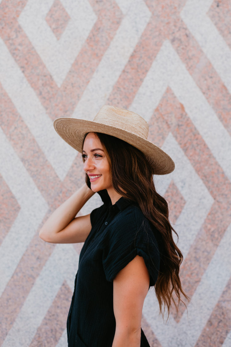Cute Summer Rompers for Women by popular Las Vegas fashion blog, Outfits and Outings: image of a woman standing next to a wall mural and wearing a black romper with a straw fedora hat. 
