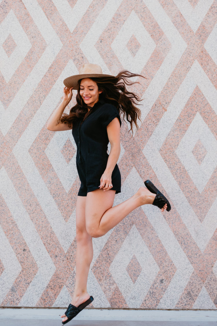 Cute Summer Rompers for Women by popular Las Vegas fashion blog, Outfits and Outings: image of a woman jumping in front of a wall mural and wearing a black romper with a straw fedora hat and black slide sandals. 
