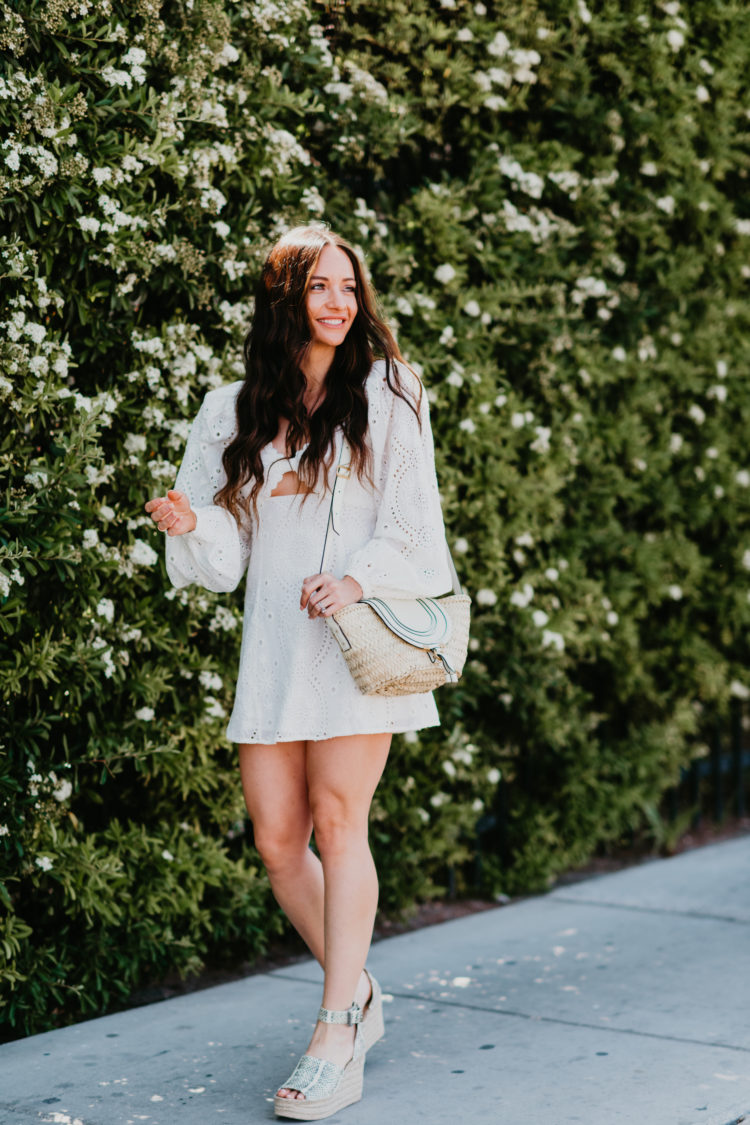 Eyelet Dresses by popular Las Vegas fashion blog, Outfits and Outings: image of a woman walking by a bush with white flowers and wearing a white eyelet dress with snake print espadrilles. 