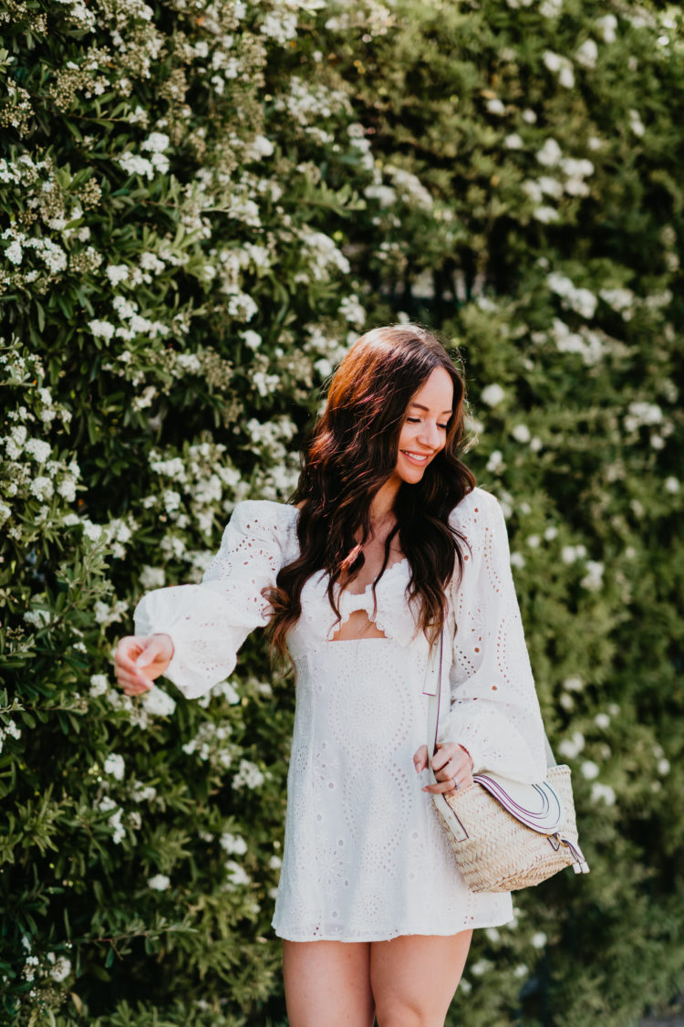 Eyelet Dresses by popular Las Vegas fashion blog, Outfits and Outings: image of a woman walking by a bush with white flowers and wearing a white eyelet dress with snake print espadrilles. 