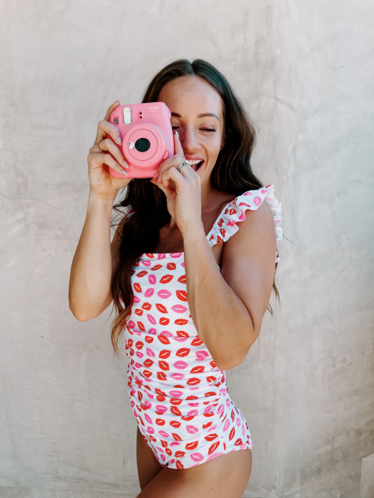 Sassy Red Lipstick by popular Las Vegas fashion blog, Outfits and Outings: image of a woman standing next to a white stucco wall and holding a pink Instax camera and wearing a white one pice swimsuit with a red and pink lips print. 